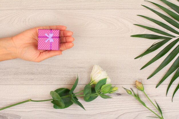 Manos de mujer sosteniendo una caja de regalo sobre fondo de madera gris con hermosas flores y hojas verdes. Concepto de dar un regalo en vacaciones o cumpleaños. Vista superior.