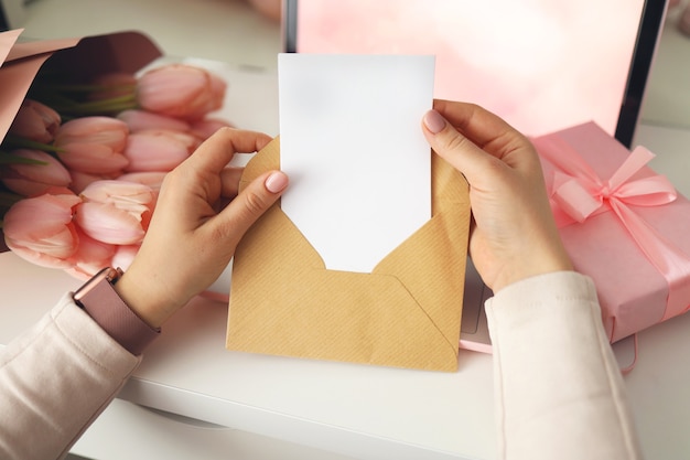 Manos de mujer sosteniendo una carta en sobre de artesanía. Fondo rosa, concepto de día de san valentín. Flor de tulipanes y caja de regalo rosa en segundo plano. Escritorio de casa para mujeres.