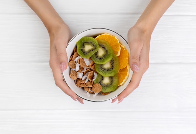 Manos de mujer sosteniendo copos multigrano con naranja y kiwi y coco en un tazón