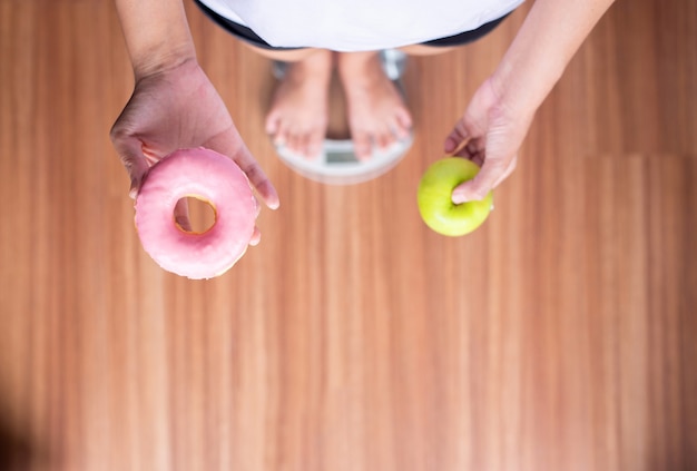 Manos de mujer sosteniendo donut rosa horneado y manzana verde durante el pie en la balanza, dieta saludable, concepto de dieta, vista superior