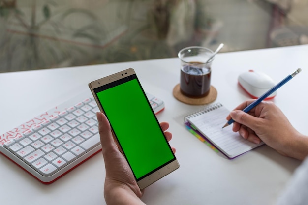 Manos de mujer sosteniendo un móvil con pantalla verde mientras bebe café Teclado y escritorio blanco