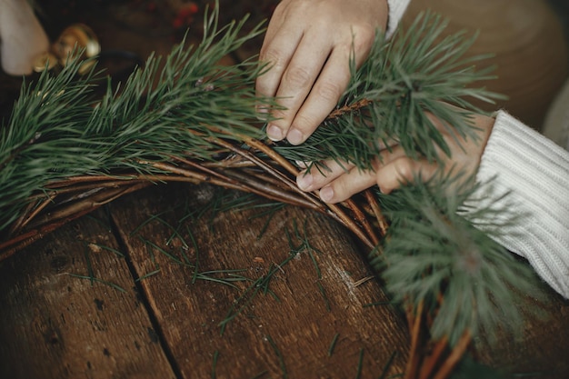 Manos de mujer sosteniendo ramas de pino arreglando corona de navidad sobre fondo rústico Taller de invierno