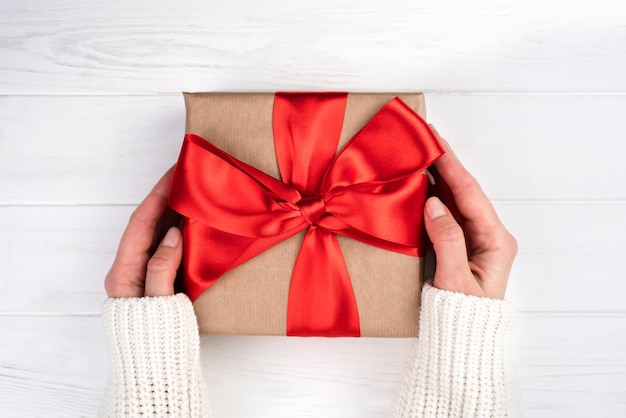 Manos de mujer sosteniendo regalo o caja de regalo con lazo rojo sobre fondo blanco de madera Primer plano Vista superior Flatlay Enfoque selectivo