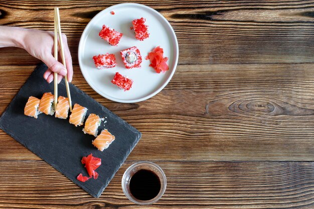 Manos de mujer sosteniendo rollos de sushi de pescado con caviar rojo salmón con palillos en plato de servir de cerámica negra gris sobre fondo rústico de madera de cerca Servicio de comida de mariscos del concepto de restaurante