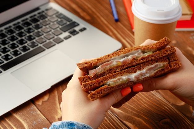 Manos de una mujer sosteniendo sándwich encima de la mesa de trabajo con portátil abierto