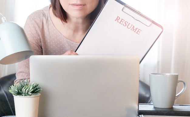Manos de mujer sosteniendo la solicitud de currículum cerca de su lugar de trabajo con computadora portátil.