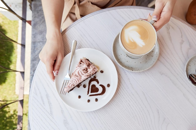 Manos de mujer sosteniendo una taza de chocolate con frutas de capuchino y pastel en una mesa de madera en la terraza del café afuera Tiempo de café atmosférico Decoraciones de postre en forma de corazón