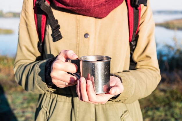 Manos de mujer sosteniendo una taza humeante en el hermoso entorno natural. Disfrutando de un café en una taza de metal turística en una excursión