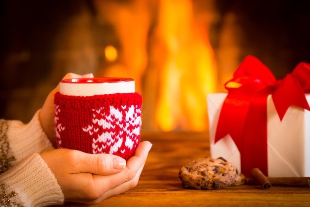 Manos de mujer sosteniendo la taza de Navidad junto a la chimenea. Concepto de vacaciones de invierno