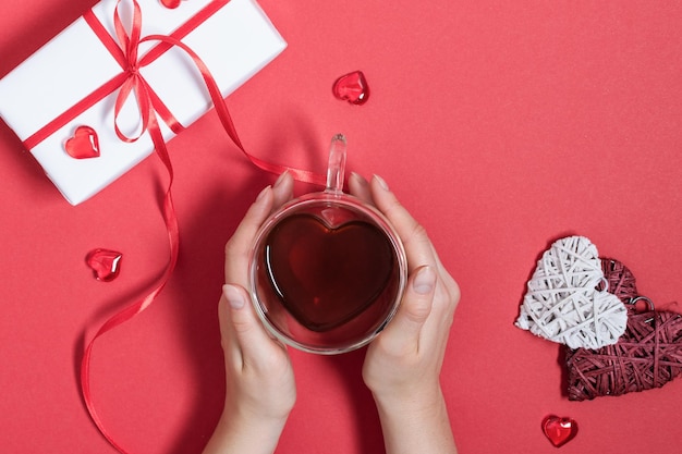 Manos de mujer sosteniendo una taza de té en forma de corazón con decoración de corazones rojos en la vista superior de la mesa roja