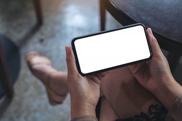 Manos de mujer sosteniendo un teléfono móvil negro con pantalla en blanco horizontalmente para ver mientras está sentado en la cafetería