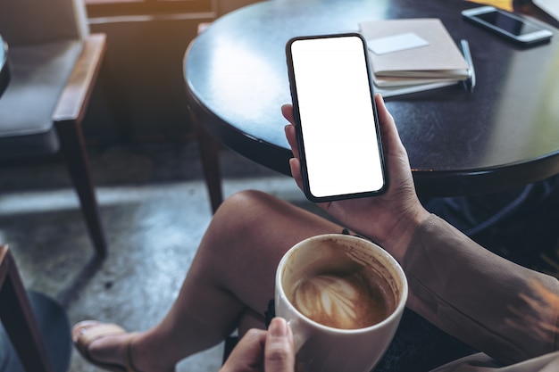 Manos de mujer sosteniendo un teléfono móvil negro con pantalla en blanco mientras bebe y se sienta en la cafetería
