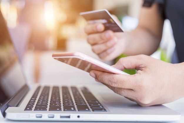 Manos de mujer sosteniendo / usando teléfono inteligente y tarjeta de crédito frente a la computadora portátil en el escritorio