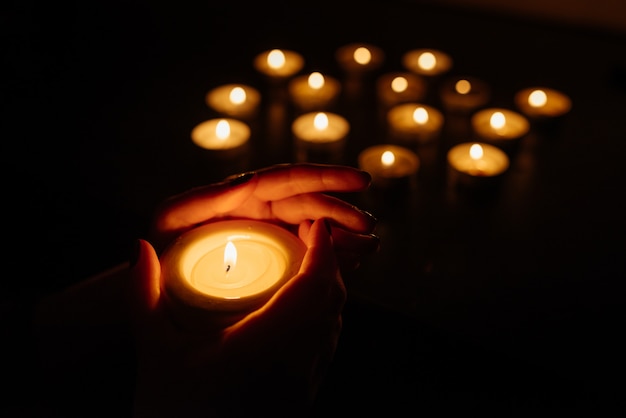 Manos de mujer sosteniendo una vela encendida. Muchas llamas de velas brillando. De cerca.