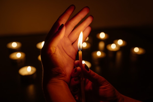 Manos de mujer sosteniendo una vela encendida. Muchas llamas de velas brillando. De cerca.