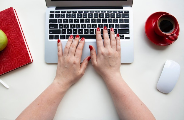 Manos de mujer en el teclado de la computadora portátil con mouse inalámbrico, taza de café, cuaderno rojo, manzana verde en la mesa blanca