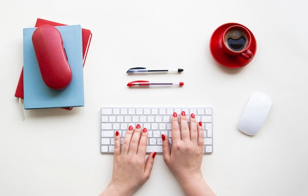 Manos de mujer en teclado y mouse blancos con bolígrafos negros y rojos, taza de café roja, cuadernos en mesa blanca