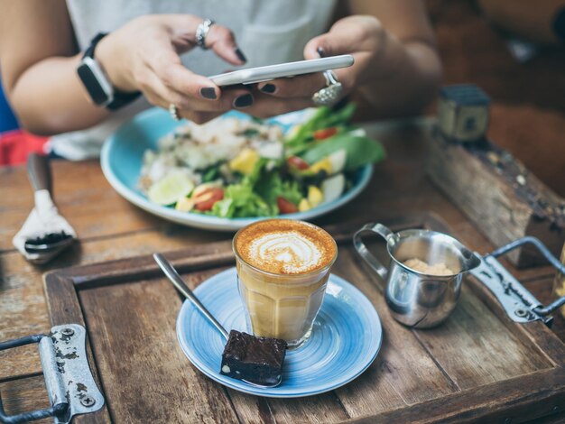 Manos de mujer tomando fotos de una taza de café en una mesa de madera con un teléfono inteligente