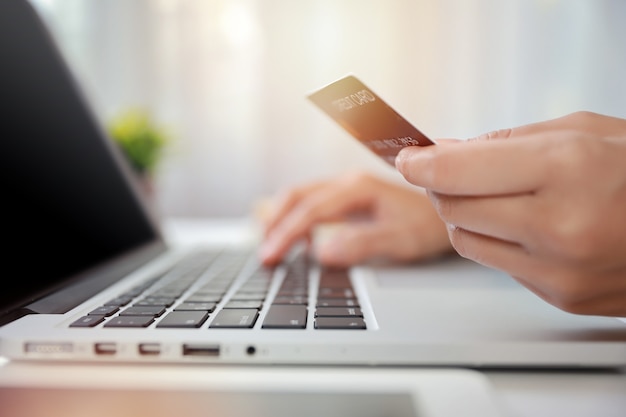 Manos de mujer en traje negro sentado y sosteniendo la tarjeta de crédito y usando la computadora portátil en la mesa para pago en línea o compras en línea. Mujer de negocios usando internet mientras trabaja. Concepto de banca electrónica