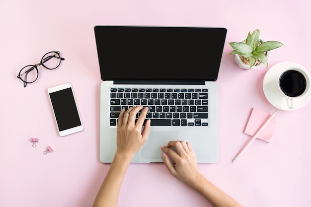 Manos de mujer usando computadora portátil con una taza de café, teléfono móvil y oficina estacionaria en rosa