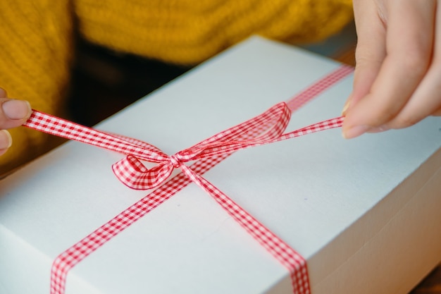 Las manos de las mujeres desatan el arco en la caja de regalo sorpresa de navidad con cinta roja a cuadros concepto de humor festivo ...