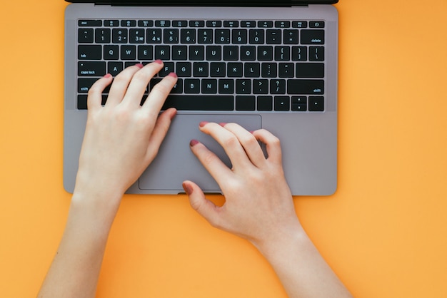 Las manos de las mujeres escriben el texto en el teclado de la computadora portátil en la superficie naranja