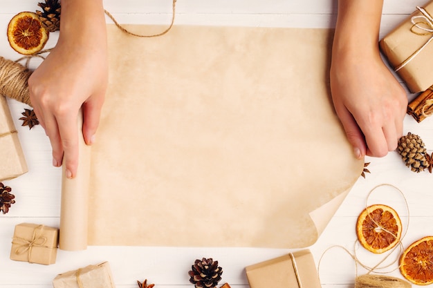 Foto las manos de las mujeres están girando el rollo de papel artesanal contra el fondo de naranja seca, canela, piñas, anís en una mesa blanca. , vista superior, lugar para el texto.