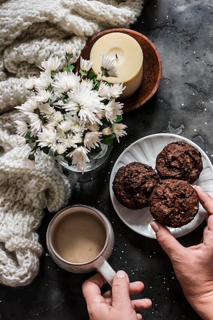 Las manos de las mujeres sostienen un pastel de chocolate y una taza de café en una mesa oscura con un acogedor ramo de bufanda de punto de crisantemos blancos y una vela en un tazón de madera Acogedor hogar