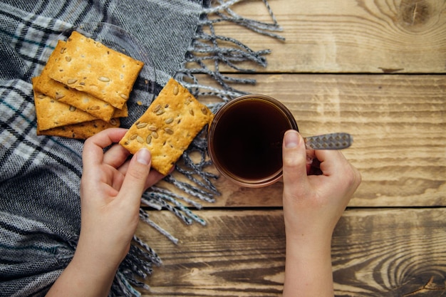 Las manos de las mujeres sostienen una taza de café negro sobre un fondo de madera. Deliciosas galletas, una cálida bufanda, un lugar para el texto.