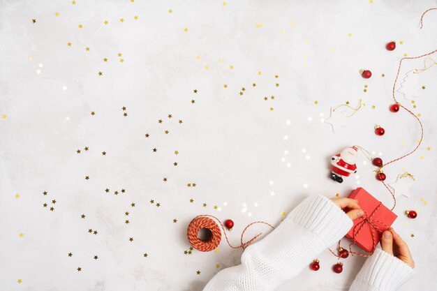 Las manos de las mujeres en un suéter llevan una caja de regalo para el año nuevo. Decoración creativa de Navidad sobre fondo blanco con confeti