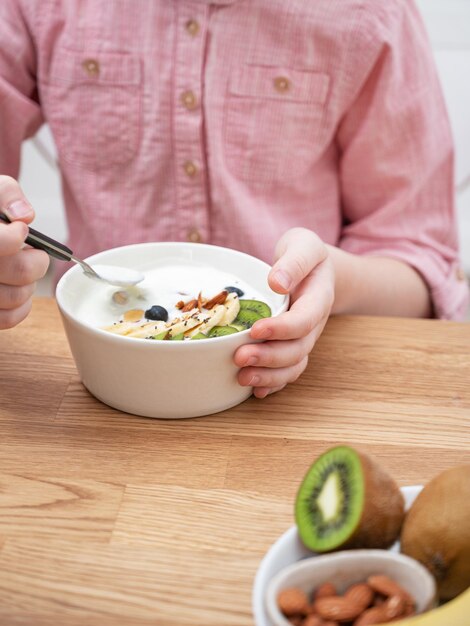 Las manos de una niña con una camisa rosa sostienen un tazón de granola de yogur y fruta fresca en una mesa de madera El concepto de alimentación saludable Orientación vertical