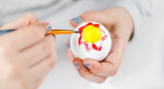 Manos de una niña caucásica pintando un huevo de Pascua con un pincel con pintura acrílica roja y amarilla