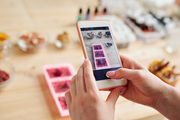 Foto manos de niña con smartphone tomando fotos de jabón artesanal en moldes de silicona sobre mesa de madera en estudio