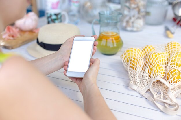 Manos de una niña sosteniendo un teléfono inteligente blanco, sobre una mesa de madera blanca, junto a limones amarillos en una bolsa