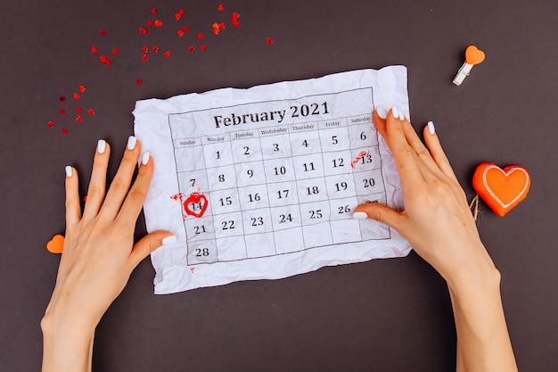 Las manos de la niña sostienen un calendario con un círculo el 14 de febrero. Vista superior. Día de San Valentín. un círculo de dulces de corazones de kars. fondo gris