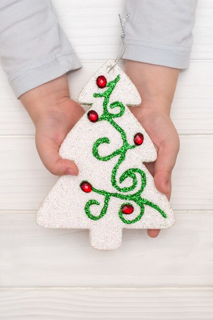 Manos del niño sosteniendo el árbol de navidad ornamental en mesa blanca