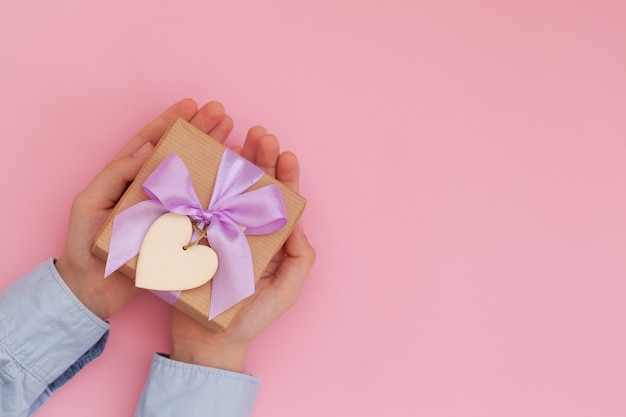 Las manos del niño sosteniendo una caja de regalo envuelta en papel artesanal y atada con un lazo en la pared rosa.