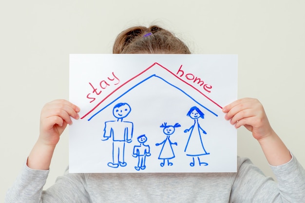 Manos de niño sosteniendo una imagen de silueta familiar bajo el techo y palabras Stay Home cubriendo su rostro sobre fondo amarillo. Niños en concepto de cuarentena.