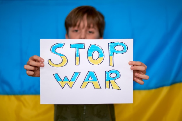 Manos de niño sosteniendo pancarta de parada de guerra dibujada en papel amarillo azul sobre fondo de bandera ucraniana