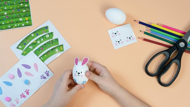 Las manos de los niños hacen una decoración de Pascua en el huevo. Lápices y pegatinas sobre un fondo amarillo. Concepto de Pascua.