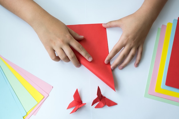 Las manos de los niños hacen origami una mariposa. El papel del color miente en una tabla.