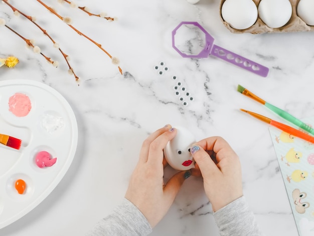 Las manos de los niños sostienen un huevo blanco y pegan ojos decorativos adhesivos sentados en una mesa de mármol