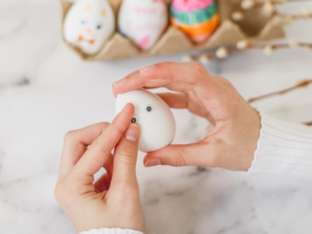 Las manos de los niños sostienen un huevo blanco y pegan pegatinas para los ojos sentados en una mesa de mármol
