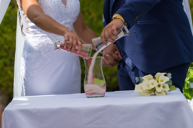 Manos del novio y la novia realizando la ceremonia de arena en un jarrón de cristal en la celebración de la boda