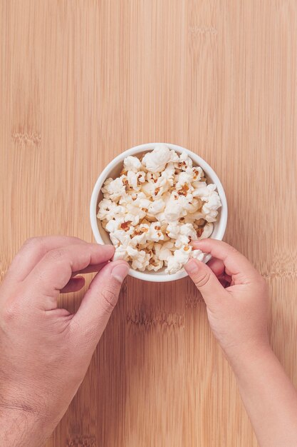 Manos de padre e hijo comiendo palomitas de maíz sobre un fondo de bambú. Planifique con el niño, vea películas con los niños, plan familiar, plan familiar.