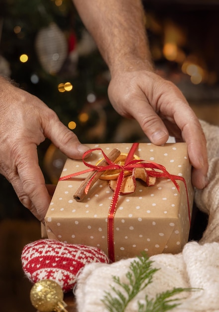 Las manos de Papá Noel ponen un regalo decorado con frutas secas contra las luces del árbol de Navidad de cerca