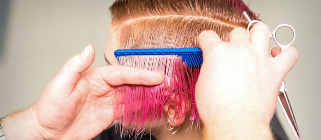 Manos de peluquero peinando el cabello haciendo peinado rosa corto para una joven mujer caucásica en un salón de belleza.