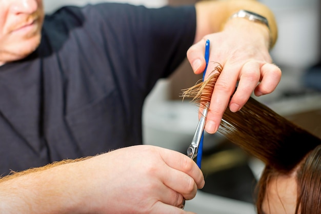 Manos de peluquero sostienen mechones de cabello entre sus dedos haciendo un corte de cabello largo de la joven con peine y tijeras en el salón de peluquería de cerca