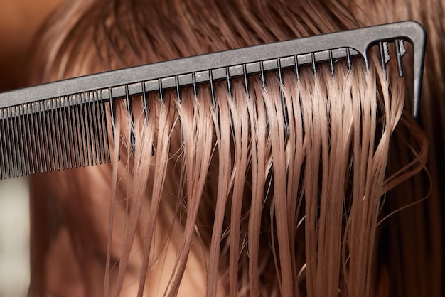 Manos de peluquero sostienen mechones de cabello entre sus dedos haciendo un corte de cabello largo de la joven con peine y tijeras en el salón de peluquería de cerca