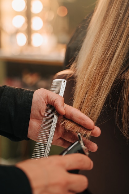 Manos de una persona irreconocible haciendo un corte de pelo con una navaja a una chica irreconocible con cabello largo y rubio. Vertical
